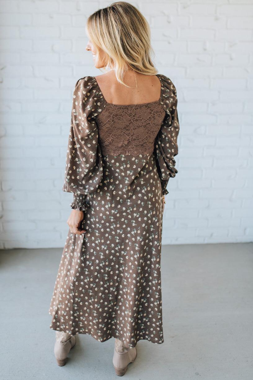 Woman wearing a brown patterned dress against a white brick wall.