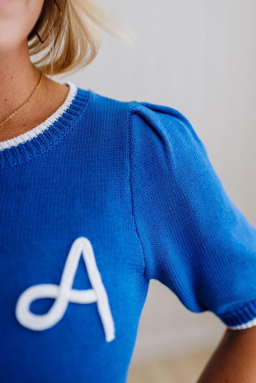 Blue sweater with a white letter 'A' on a neutral background
