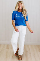 Woman wearing a blue top with 'USA' text and white pants, standing against a light wooden panel background.