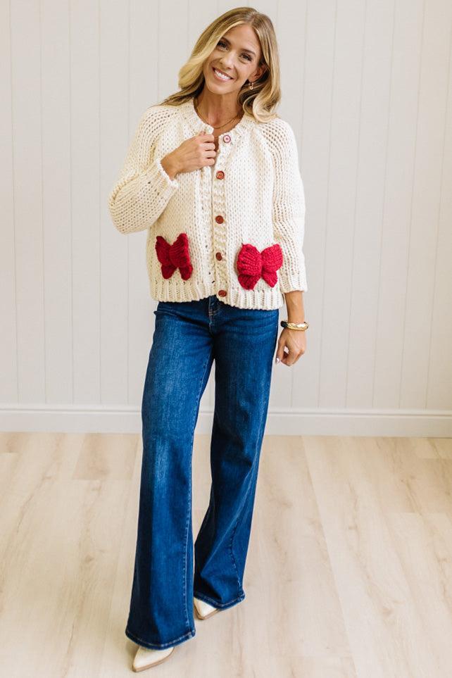 Blonde Woman in a Bright Studio wearing a cream Front Button Closure chunky sweater with red Bow Trim, Long Sleeves.