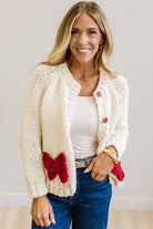 Blonde Woman in a Bright Studio wearing a cream Front Button Closure chunky sweater with red Bow Trim, Long Sleeves.