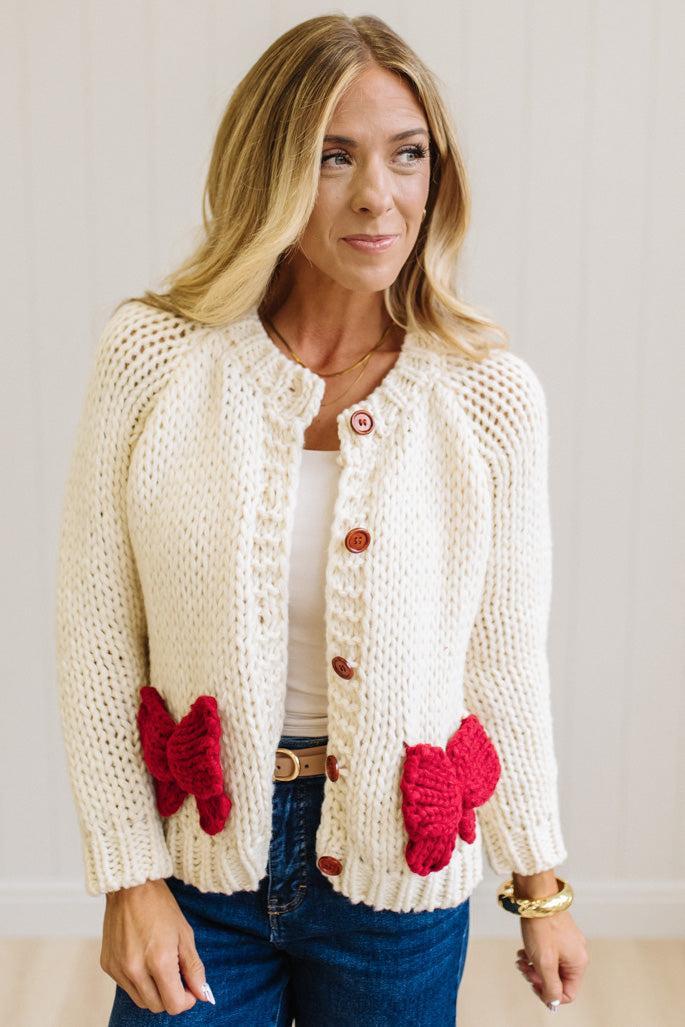 Blonde Woman in a Bright Studio wearing a cream Front Button Closure chunky sweater with red Bow Trim, Long Sleeves.