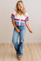 A woman modeling a white knit short-sleeve top with red and blue striped details, paired with blue jeans and tan sandals, standing against a light wood-paneled wall.
