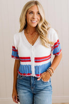 A woman wearing a white knit cardigan with red and blue striped details on the sleeves and hem, paired with blue jeans, standing against a light beige paneled wall.