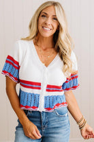 A smiling blonde woman wearing a white knit cardigan with red and blue striped details on the sleeves and hem, paired with blue jeans.