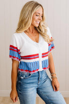 A woman wearing a white knit cardigan with blue and red striped details on the sleeves and hem, paired with blue jeans, standing against a light beige paneled wall.