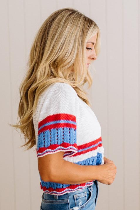 A woman shown from the side profile wearing a white knit top with red and blue horizontal striped patterns, paired with blue jeans, against a light beige paneled wall.