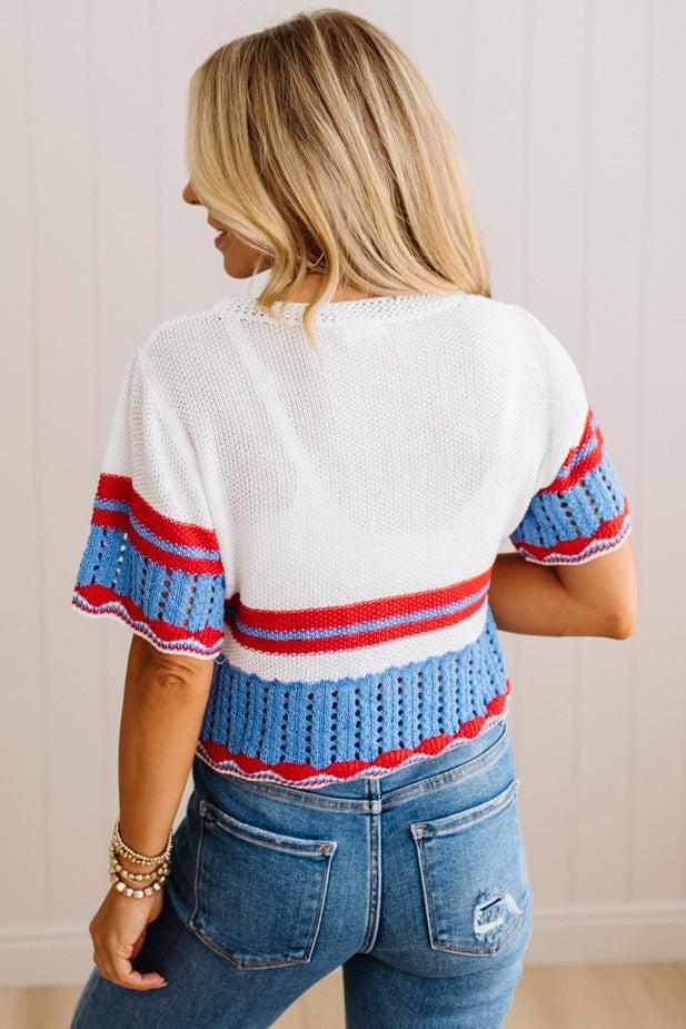 A woman shown from the back wearing a white knit top with blue and red striped details on the sleeves and hem, paired with blue jeans, set against a light beige paneled wall.
