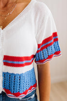 A close-up of a person wearing a white knit short-sleeve top with red and blue striped accents, paired with blue jeans and layered gold necklaces.