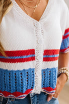 A close-up of a woman wearing a white knit cardigan with red and blue horizontal stripes, paired with blue jeans and layered gold necklaces.