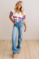 A woman modeling a white short-sleeve knit top with red and blue striped details on the sleeves and hem, paired with light-wash blue jeans and tan sandals, standing against a white paneled wall and light wood floor.