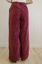 Maroon corduroy pants worn by a person on a light wooden floor with a white paneled wall background.