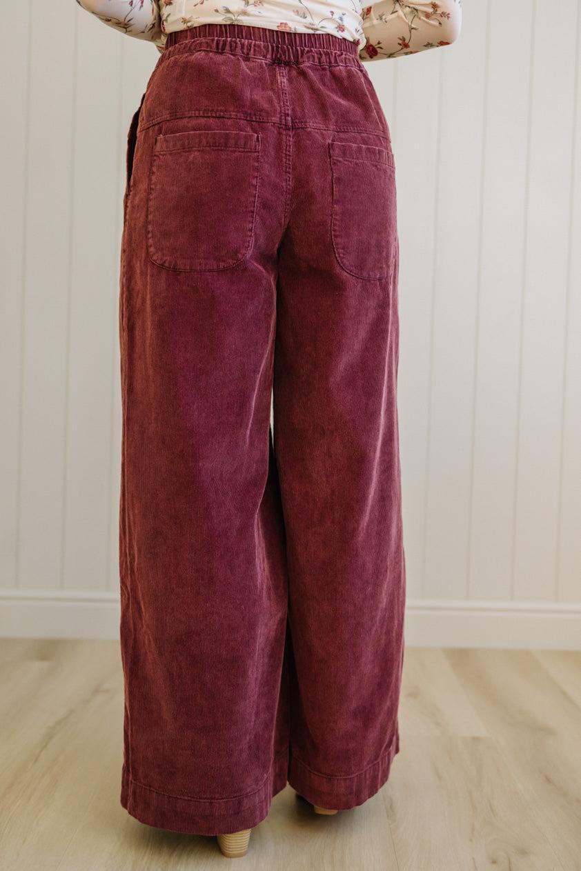 Maroon corduroy pants worn by a person on a light wooden floor with a white paneled wall background.