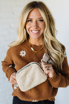 Woman wearing a brown sweater with floral embellishments, holding a beige pouch and phone.