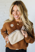 Woman wearing a brown sweater with floral patterns, holding a beige pouch and phone.