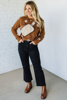 Woman wearing a brown sweater with a beige bag, black pants, and brown shoes against a white brick wall.