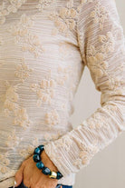 Blonde Woman Wearing A Floral Embroidered Stretch Lace Long Sleeve Top with Dark Jeans and Light Brown Belt.