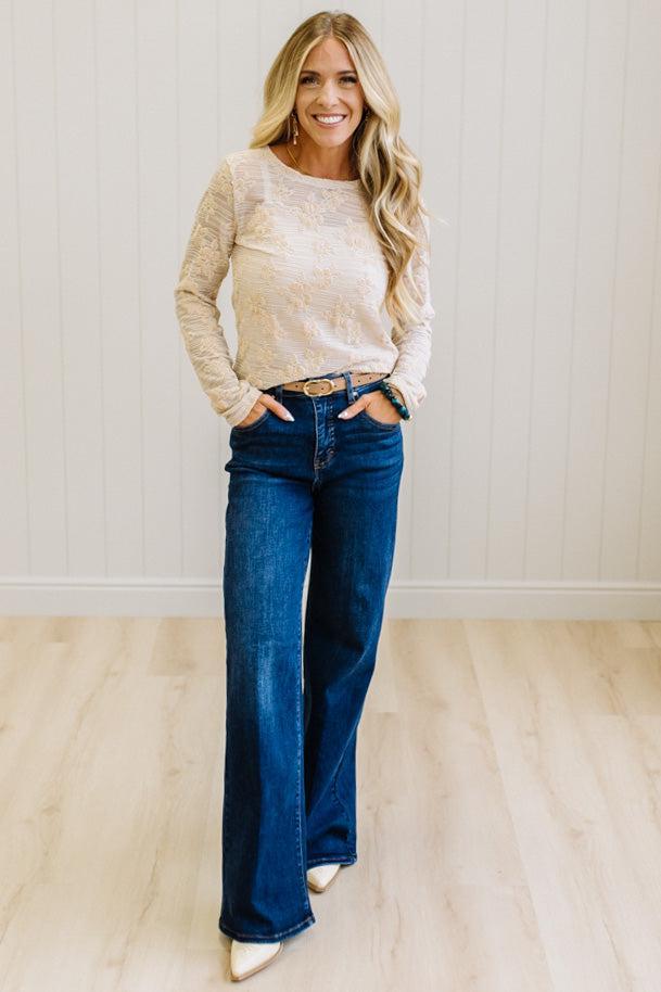 Blonde Woman Wearing A Floral Embroidered Stretch Lace Long Sleeve Top with Dark Jeans and Light Brown Belt.