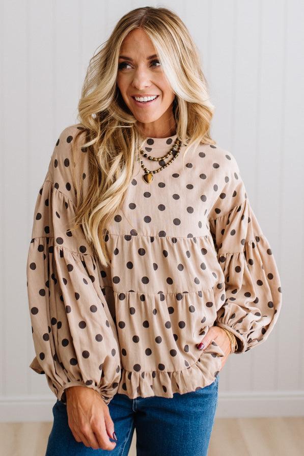 BLONDE WOMAN WEARING A WHIMSICAL POLKADOT RUFFLE PUFF BLOUSE IN LATTE STANDING AGAINST A PANELED WALL IN A BRIGHTLY LIT STUDIO.