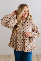 BLONDE WOMAN WEARING A WHIMSICAL POLKADOT RUFFLE PUFF BLOUSE IN LATTE STANDING AGAINST A PANELED WALL IN A BRIGHTLY LIT STUDIO.