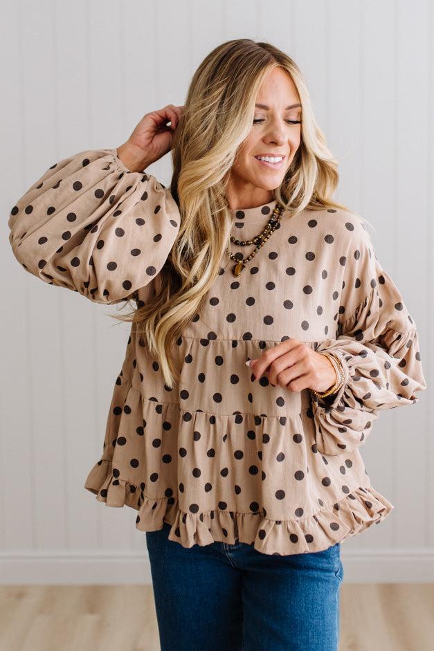 BLONDE WOMAN WEARING A WHIMSICAL POLKADOT RUFFLE PUFF BLOUSE IN LATTE STANDING AGAINST A PANELED WALL IN A BRIGHTLY LIT STUDIO.