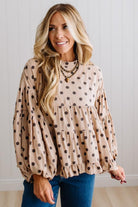 BLONDE WOMAN WEARING A WHIMSICAL POLKADOT RUFFLE PUFF BLOUSE IN LATTE STANDING AGAINST A PANELED WALL IN A BRIGHTLY LIT STUDIO.