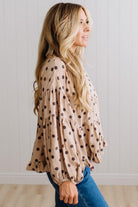 BLONDE WOMAN WEARING A WHIMSICAL POLKADOT RUFFLE PUFF BLOUSE IN LATTE STANDING AGAINST A PANELED WALL IN A BRIGHTLY LIT STUDIO.