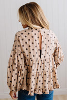BLONDE WOMAN WEARING A WHIMSICAL POLKADOT RUFFLE PUFF BLOUSE IN LATTE STANDING AGAINST A PANELED WALL IN A BRIGHTLY LIT STUDIO.