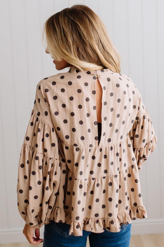 BLONDE WOMAN WEARING A WHIMSICAL POLKADOT RUFFLE PUFF BLOUSE IN LATTE STANDING AGAINST A PANELED WALL IN A BRIGHTLY LIT STUDIO.