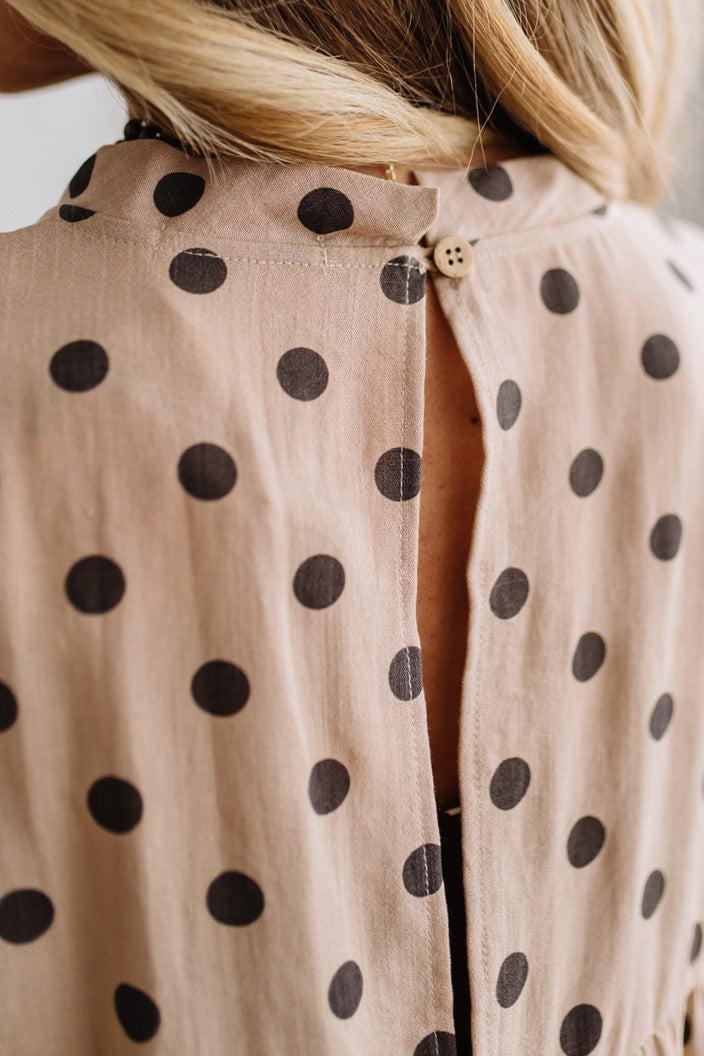 BLONDE WOMAN WEARING A WHIMSICAL POLKADOT RUFFLE PUFF BLOUSE IN LATTE STANDING AGAINST A PANELED WALL IN A BRIGHTLY LIT STUDIO.
