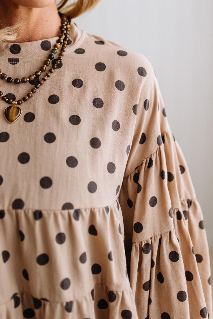 BLONDE WOMAN WEARING A WHIMSICAL POLKADOT RUFFLE PUFF BLOUSE IN LATTE STANDING AGAINST A PANELED WALL IN A BRIGHTLY LIT STUDIO.