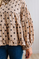 BLONDE WOMAN WEARING A WHIMSICAL POLKADOT RUFFLE PUFF BLOUSE IN LATTE STANDING AGAINST A PANELED WALL IN A BRIGHTLY LIT STUDIO.