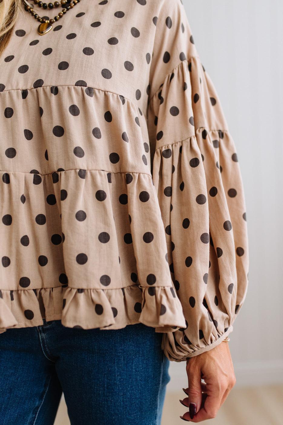 BLONDE WOMAN WEARING A WHIMSICAL POLKADOT RUFFLE PUFF BLOUSE IN LATTE STANDING AGAINST A PANELED WALL IN A BRIGHTLY LIT STUDIO.