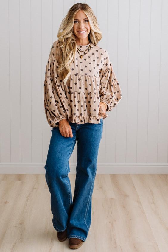 BLONDE WOMAN WEARING A WHIMSICAL POLKADOT RUFFLE PUFF BLOUSE IN LATTE STANDING AGAINST A PANELED WALL IN A BRIGHTLY LIT STUDIO.