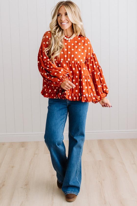 BLONDE WOMAN WEARING A WHIMSICAL POLKADOT RUFFLE PUFF BLOUSE IN RUST STANDING AGAINST A PANELED WALL IN A BRIGHTLY LIT STUDIO.
