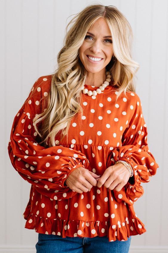 BLONDE WOMAN WEARING A WHIMSICAL POLKADOT RUFFLE PUFF BLOUSE IN RUST STANDING AGAINST A PANELED WALL IN A BRIGHTLY LIT STUDIO.