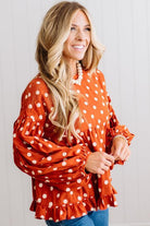 BLONDE WOMAN WEARING A WHIMSICAL POLKADOT RUFFLE PUFF BLOUSE IN RUST STANDING AGAINST A PANELED WALL IN A BRIGHTLY LIT STUDIO.