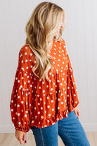 BLONDE WOMAN WEARING A WHIMSICAL POLKADOT RUFFLE PUFF BLOUSE IN RUST STANDING AGAINST A PANELED WALL IN A BRIGHTLY LIT STUDIO.