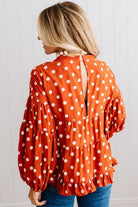BLONDE WOMAN WEARING A WHIMSICAL POLKADOT RUFFLE PUFF BLOUSE IN RUST STANDING AGAINST A PANELED WALL IN A BRIGHTLY LIT STUDIO.
