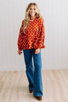 BLONDE WOMAN WEARING A WHIMSICAL POLKADOT RUFFLE PUFF BLOUSE IN RUST STANDING AGAINST A PANELED WALL IN A BRIGHTLY LIT STUDIO.