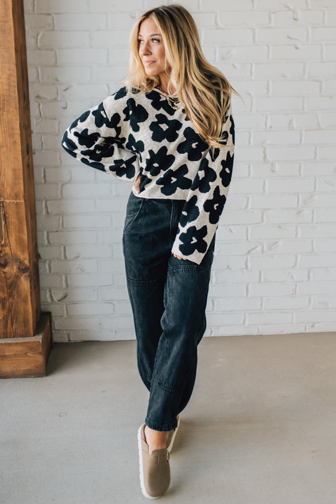 Woman wearing a floral sweater and dark jeans against a white brick wall.