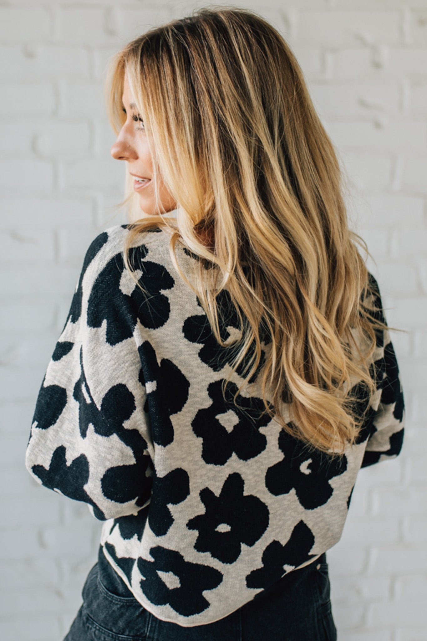 Woman wearing a black and white floral patterned sweater against a white brick wall.