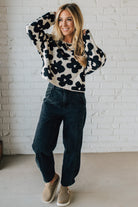 Woman wearing a floral sweater and dark jeans against a white brick wall.