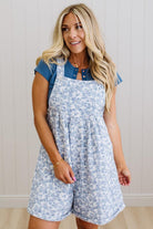 Woman wearing a blue floral overalls dress with a denim shirt underneath against a light background