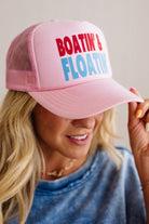 Pink trucker hat with 'Boatin' & Floatin'' text held by a person wearing a blue shirt.