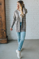 Woman wearing a striped cardigan, white shirt, and light blue jeans against a white brick wall.