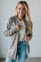 Woman wearing a long, striped cardigan over a white shirt and blue jeans against a white brick wall.