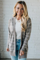 Woman wearing a gray striped cardigan over a white shirt and blue jeans, standing against a white brick wall.