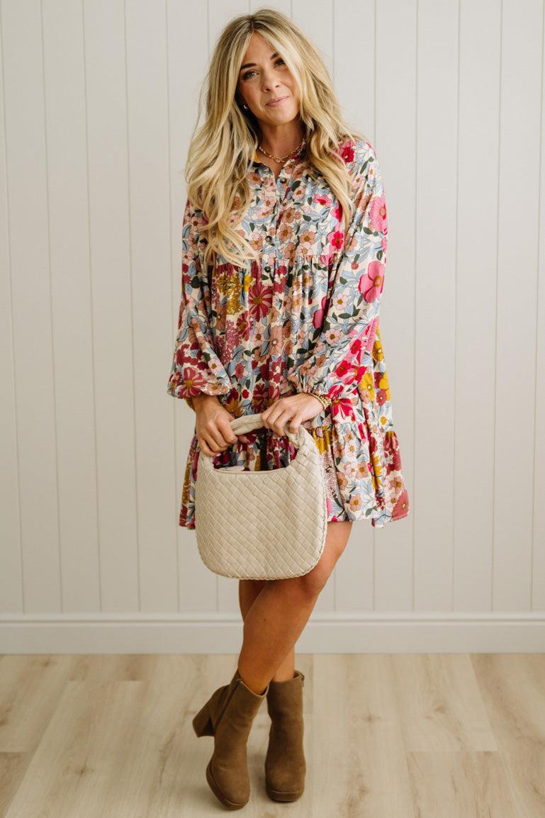 Woman wearing a floral dress holding a beige bag against a white paneled wall.