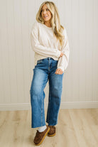 Woman wearing a white sweater, blue jeans, and brown shoes standing against a white paneled wall.
